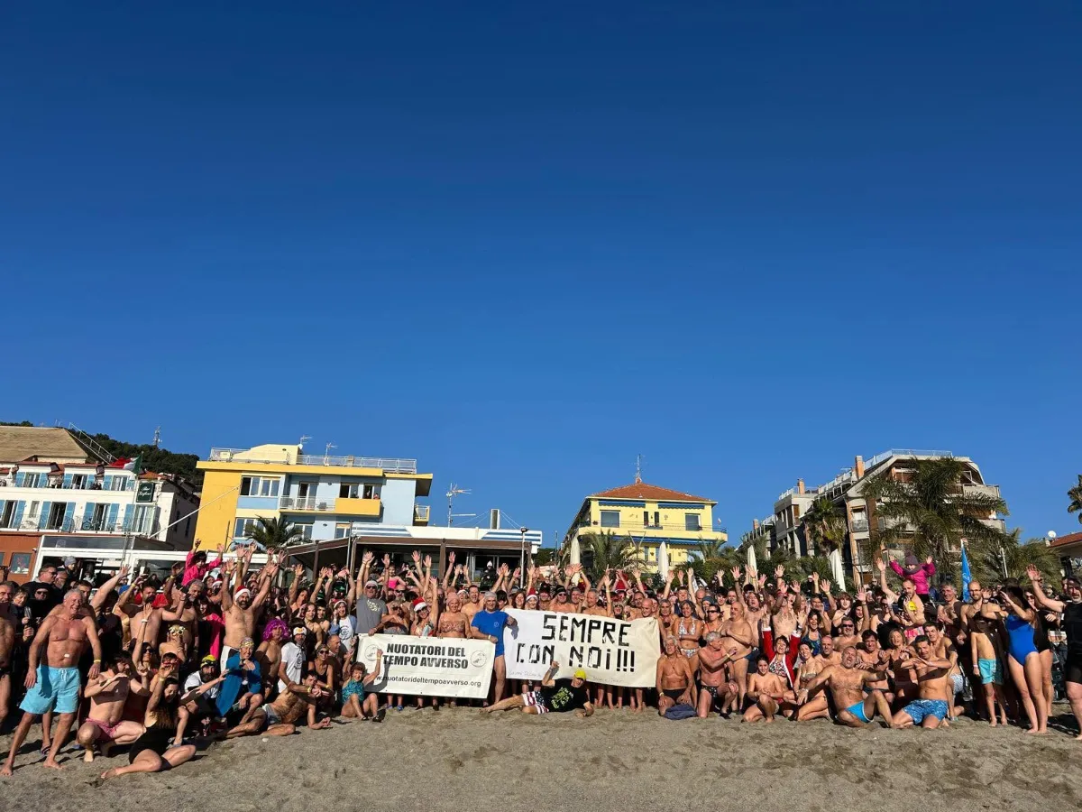 Numeroso gruppo di persone in costume sulla spiaggia, con striscioni e mani alzate.