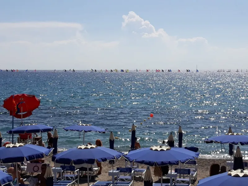 Ombrelloni blu e lettini sulla spiaggia, con numerose barche a vela all’orizzonte.