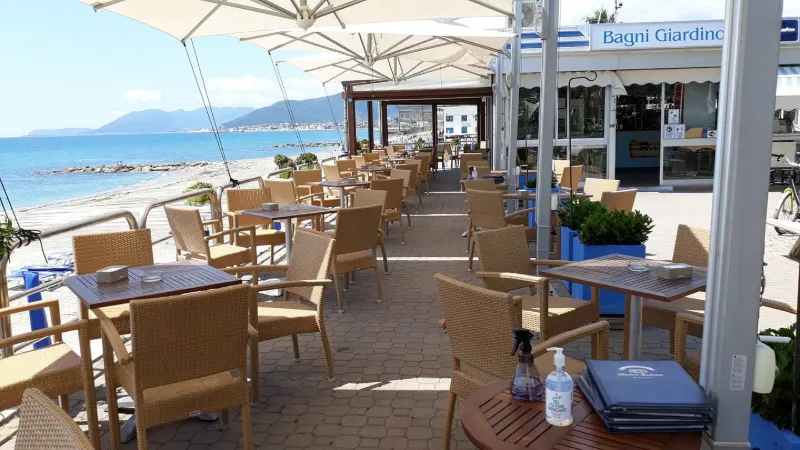 Terrazza sul mare con tavoli, sedie in vimini e grandi ombrelloni bianchi.