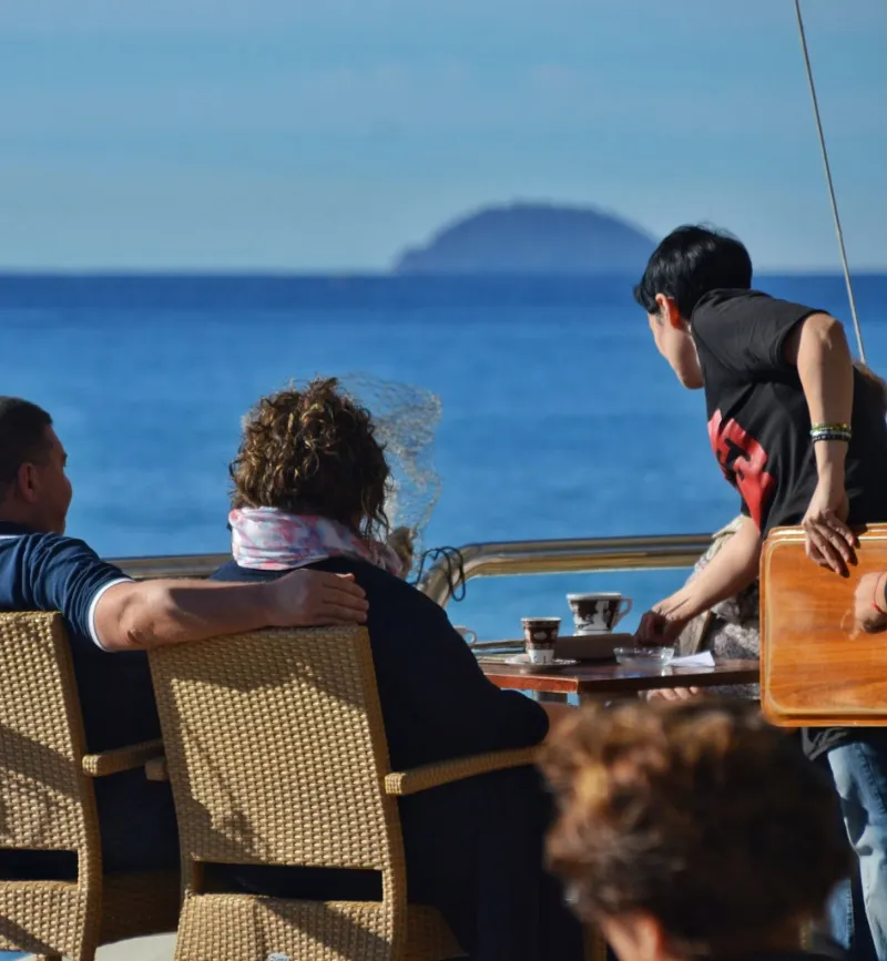 Persone sedute al tavolino con tazzine su una terrazza affacciata sul mare, un’isola sullo sfondo.