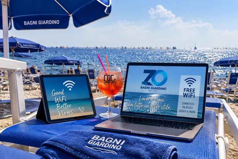 Computer portatile, tablet e cocktail su lettino, con indicazioni Wi-Fi e spiaggia sullo sfondo.